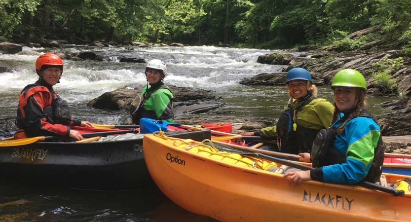 People wearing safety gear sit in canoes and smile at the camera. In the background, there is whitewater and trees lining the shores.